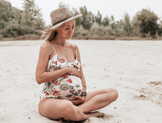 Young happy pregnant woman in a swimsuit and hat sitting on sand. pregnancy concept