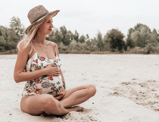 Young happy pregnant woman in a swimsuit and hat sitting on sand. pregnancy concept