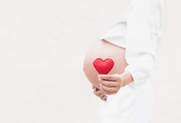 Young woman hand holding red bright heart shape on naked belly. Emotional loving moment in pregnancy time - 37 weeks. Baby expectation. Love, happiness and safety concept. Closeup. White background