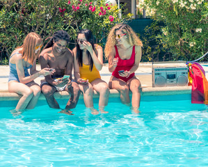 Group of people of different ethnic groups with bathing suits of different colors with the mobile in the hand sitting on the edge of a swimming pool with a cassette and a lgtb flag