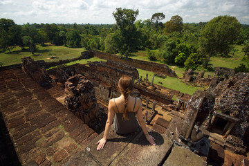 Obraz premium Young woman sitting on stones of old Angkor temple on background of bright green landscape and ancient ruins, Cambodia