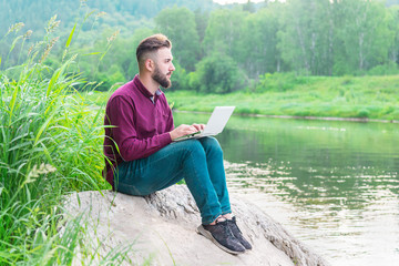 Fototapeta premium A young man in a shirt and jeans sits on a stone against the background of nature near the river on a summer day with a laptop. remote work and freelance concept