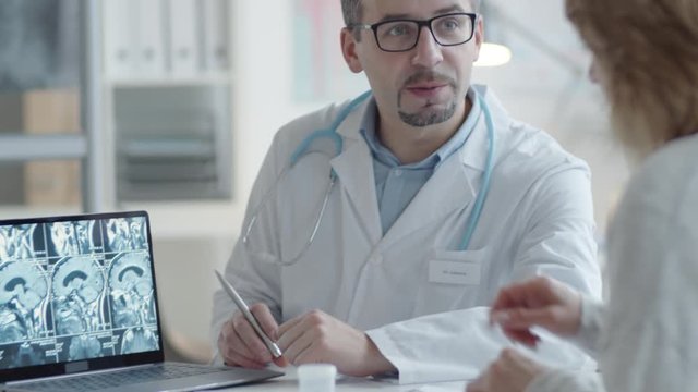 Tilt up shot of caucasian male doctor zooming and explaining x-ray image of brain on laptop screen to female patient while speaking in medical office