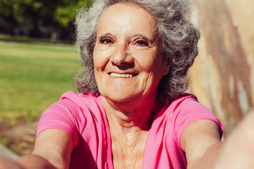 Happy old lady taking selfie while walking in park. Self portrait of senior grey haired woman in casual with grass and trees in background. Self shot concept