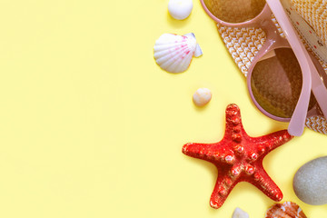 Top view of sunglasses, straw hat, sunglasses and shells with a red starfish and sea pebbles. Vacation background. Summertime, travel, beach, tourism concept. Yellow background, copy space.