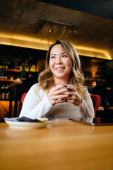 Beautiful Asian woman drinking coffee in warm cosy coffee shop and looking away	