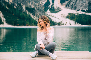 Fototapeta na wymiar Beautiful girl long flowing hair sits on wooden pier, legs lotus position, hand near head smiles enjoy nature relax silence beauty of alpine Lake Braies. Backdrop white mountains green forest water