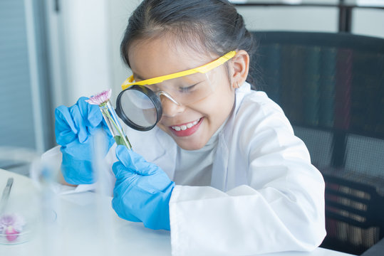 Little Asian Girl Elementary School Looking Into Magnifying Glass At The Flower On Desk At School.scientist Making Experiments In Home Laboratory. Child And Science. Education Concept. .