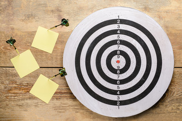 Dartboard with darts and paper sheets on wooden background