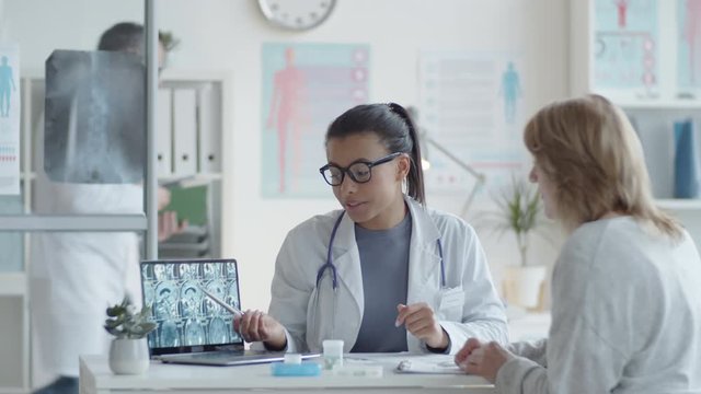 Young beautiful mixed raced female doctor showing x-ray image of brain on laptop screen and answering questions from patient during medical consultation in clinic