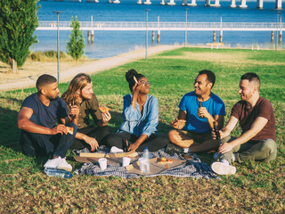 Cheerful smiling friends having picnic in park. Young people sitting on green grass and eating...