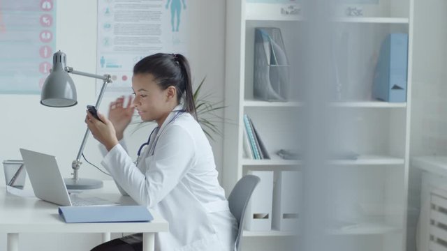 Side view of young mixed raced female doctor in lab coat sitting at desk with laptop on it and using smartphone while working in medical office