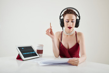 Beautiful female singer in headphones reading lyrics in document and practicing new song