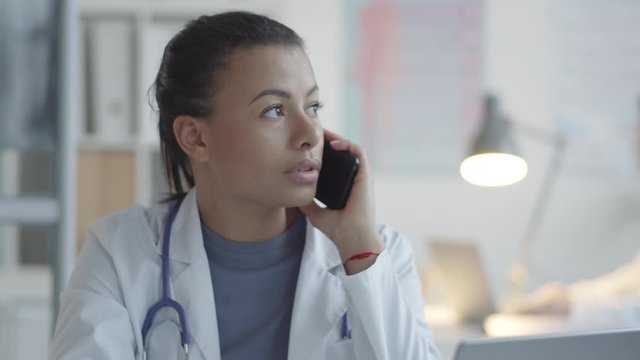 Tilt up shot of attractive mixed raced female doctor in lab coat with stethoscope over neck talking on mobile phone while working at desk in medical office