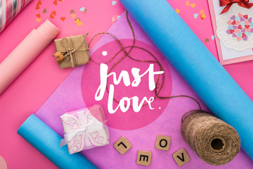 top view of valentines decoration, wrapping paper, twine, gift boxes, greeting card and love lettering on wooden cubes on pink background with just love lettering