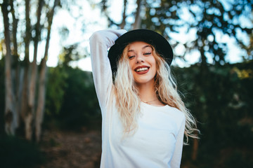 Chica feliz rubia sujetando su sombrero con fondo natural verde.