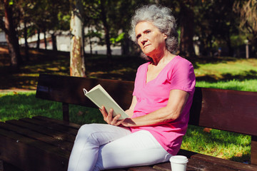 Serious focused old lady reading novel in park. Senior grey haired woman in casual sitting on...