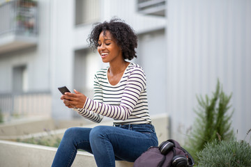 Obraz premium smiling young black woman sitting outside looking at mobile phone