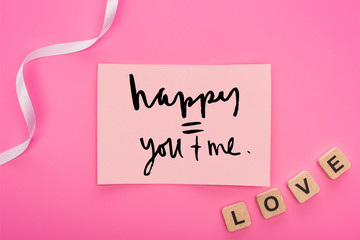 top view of empty paper card with happy you and me lettering near ribbon and love lettering on wooden cubes isolated on pink