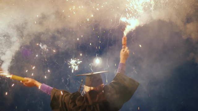 An emotional college graduate in a mantle and a graduation cap with two fireworks in his hands