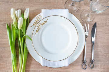 Beautiful decorated table setting. White plates, forged fork and knife, set of wineglasses and bouquet of white tulips on wooden background. Top view