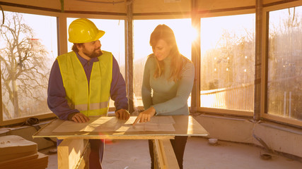 LENS FLARE: Autumn sunbeams shine on woman and contractor discussing floor plans - obrazy, fototapety, plakaty
