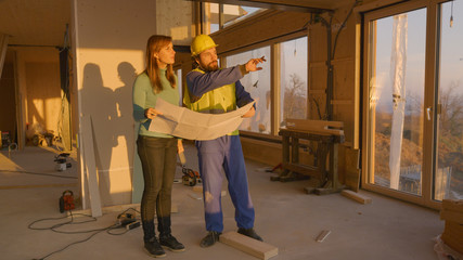 Contractor in overalls shows home owner around the unfinished house at sunset