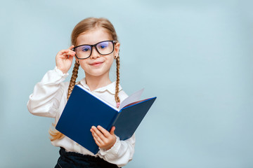 Cheerful young blonde child girl with glasses holding a book to study isolated on a blue background