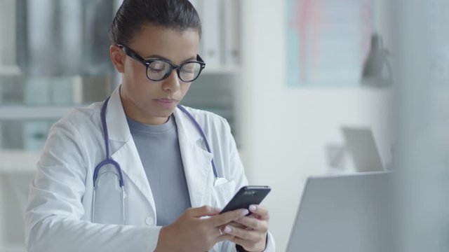 Beautiful mixed raced female doctor in lab coat and glasses sitting at desk in medical office and scrolling on smartphone screen while browsing the Internet
