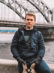 Portrait of a blond Man in a jacket and sweater with a backpack stands on the background of the river with a bridge, on the bridge busy traffic