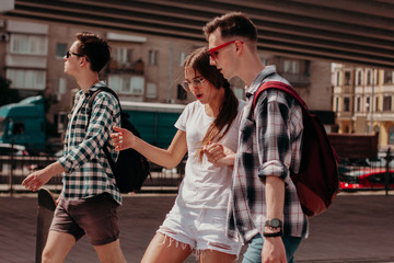 Friends Students Walk Around The City During The Summer Holidays. Guys In Plaid Shirts And A Girl In White Walk Along A Busy Street Of A European City. The Girl Dances On The Run.