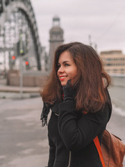 Portrait of a beautiful brunette girl with long hair in a coat and with a bright scarf, stands against the background of the river with a bridge, on the bridge busy traffic