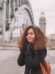 Portrait of a beautiful brunette girl with long hair in a coat and with a bright scarf, stands against the background of the river with a bridge, on the bridge busy traffic