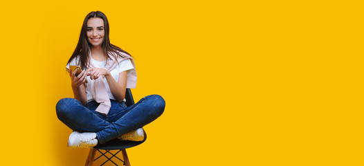 Charming caucasian girl sitting on a chair and using a phone while posing near yellow freespace