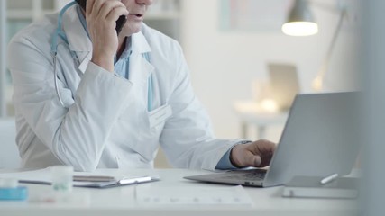 Midsection shot of senior male doctor using laptop and talking on mobile phone while working at desk in clinic - Powered by Adobe