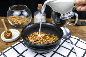 Pouring milk into black bowl of homemade granola with nuts, oat flakes, raisins. Healthy food concept. Fitness breakfast