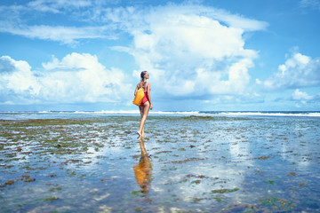 Tropical traveling. Young woman with waterproof backpack walking by low tide sea beach.