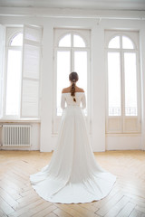 Beautiful slender blonde in the evening sun in a long white dress near a large window. Perfect hair and makeup for the bride, a new collection of wedding dresses