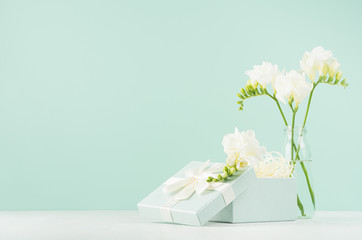 Spring gentle celebration background with opened gift box with paper filler, flowers in glass vase in elegant green mint menthe interior on white  board.