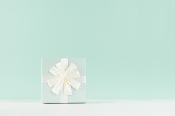 Elegant delicate celebration background with closed standing square gift box with ribbon and knot on green mint menthe interior on white wood table.
