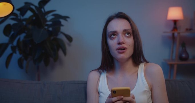 Close up of young girl crying while looking photos with her boyfriend after broke up. Sad young woman sitting on sofa looking at smartphone screen and weeping after receiving bad news .