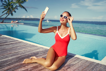 Vacation and technology. Colorful portrait of pretty young woman taking selfie portrait with her  smartphone near swimming pool on tropical beach.