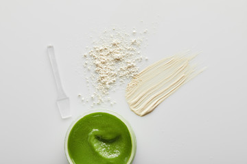 Top view of container with scrub, tone cream brush strokes, face powder and spatula on grey background