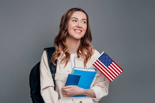 Happy girl student holding backpack, book, notebook, passport and USA flag isolated on a dark grey background, copy space
