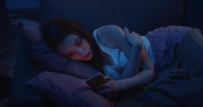 View from above of young sleepless woman lying on sofa and starring at phone screen . Depressed millennial girl missing her ex boyfriend after brokeup and looking at their photos at smartphone.