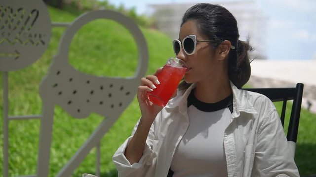 cute Asian girl wearing white shirt sitting at table outdoor, drinking red refresher through a straw, looking away and smiling, a wonderful summer day, copy space