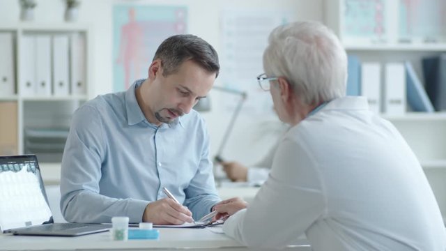 Young Caucasian man signing health service agreement and giving handshake to senior doctor while visiting medical office