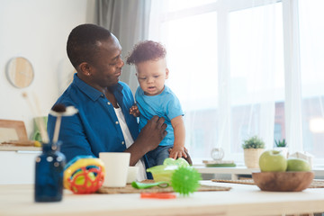 Portrait of happy African-American dad holding cute little daughter white sitting at table with...