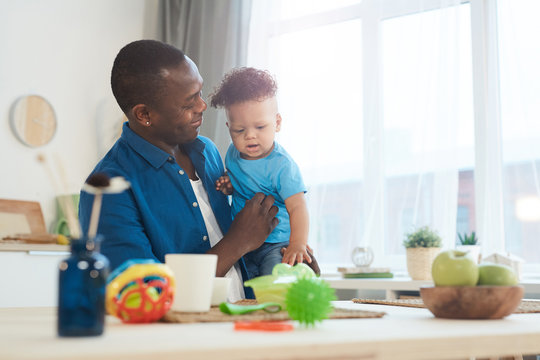 Portrait of happy African-American dad holding cute little daughter white sitting at table with children toys, copy space