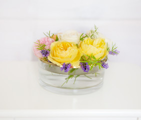 Yellow, pink and white roses in a glass vase on a background of a wooden white wall